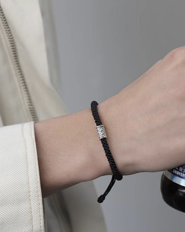 A person wearing a black braided bracelet with a silver charm, dressed in a light-colored jacket.