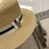 A close-up of a beige straw boater hat, highlighting the black-and-white ribbon band with a silver tree-shaped charm.