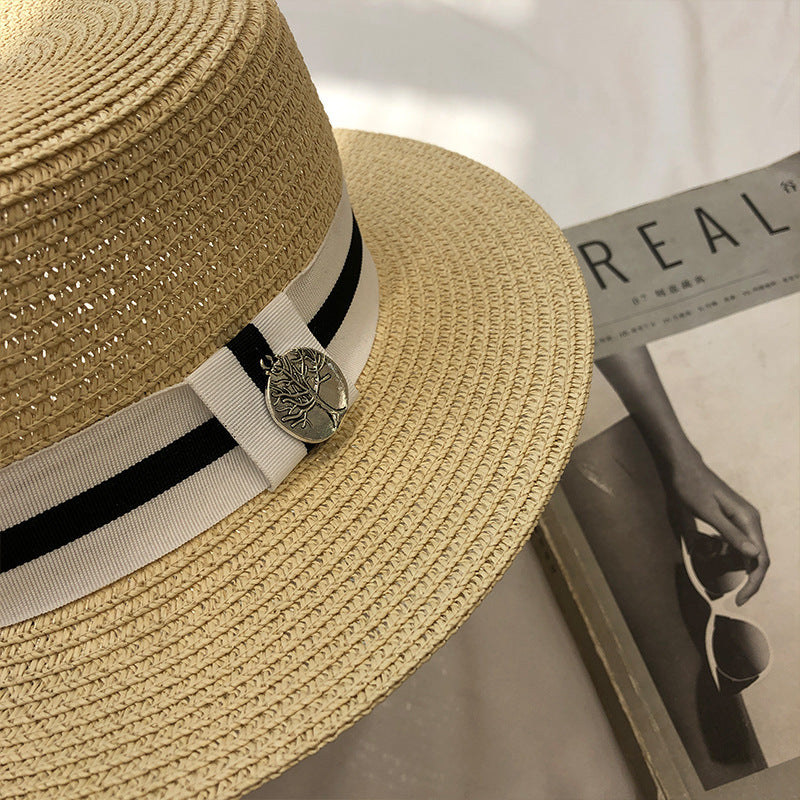 A close-up of a beige straw boater hat, highlighting the black-and-white ribbon band with a silver tree-shaped charm.