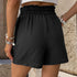 Detail shot of black paperbag waist shorts showing textured fabric and elastic gathering, styled with silver charm bracelets