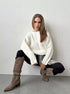 Cream oversized ribbed sweater styled with black pants, plaid skirt detail, and taupe knee-high cowboy boots, against white backdrop