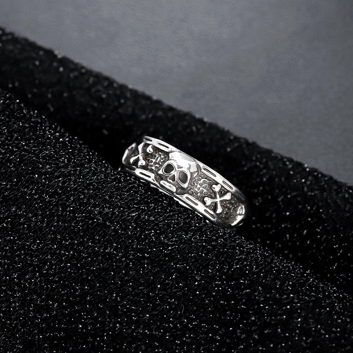A silver skull ring with crossbones details resting on a black textured surface, emphasizing its rugged and gothic aesthetic.