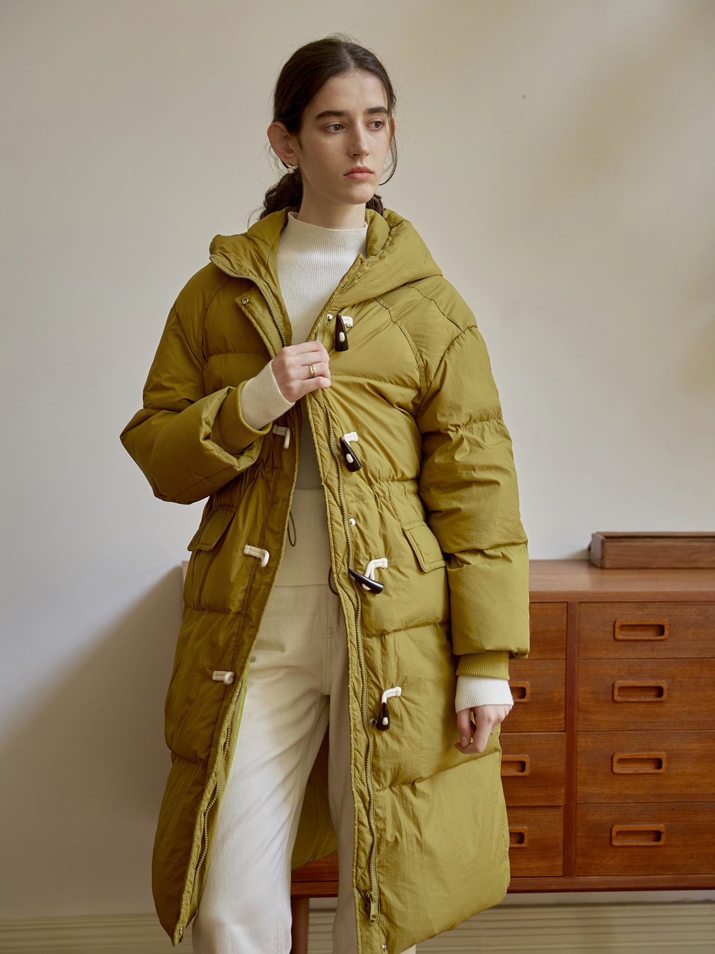 Long olive down coat featuring white toggle closures and utility pockets, styled with cream knitwear