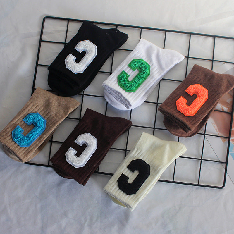 Six pairs of folded varsity-style crew socks in black, cream, brown, beige, and white with colorful "C" patches are neatly placed on a wire grid
