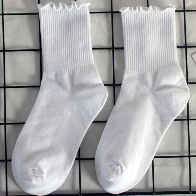 A pair of white cotton ankle socks with ruffled trim edge placed on a black grid display, showing detailed ribbed texture and cuffs