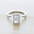 A silver solitaire engagement ring with emerald-cut diamond center stone and thin pavé diamond band, resting on soft white surface