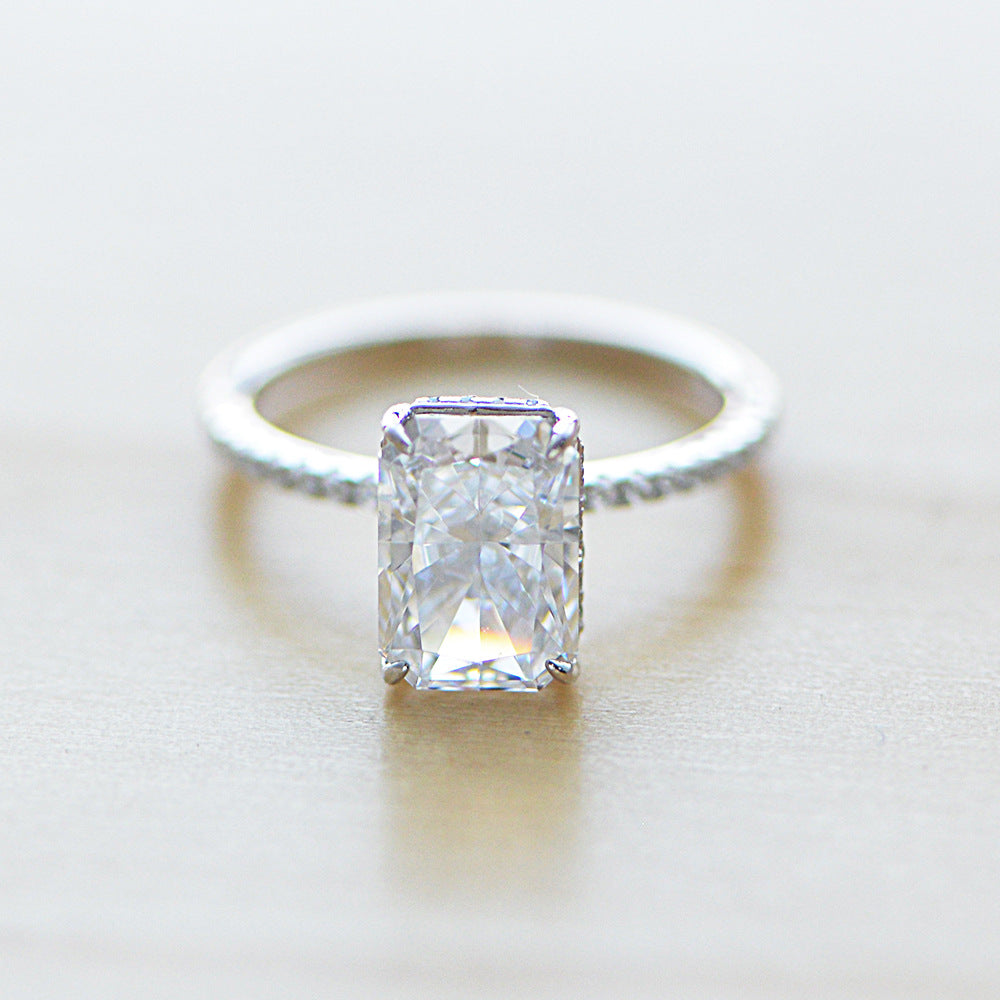 A silver solitaire engagement ring with emerald-cut diamond center stone and thin pavé diamond band, resting on soft white surface
