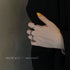 A woman's hand with yellow nail polish wearing three silver rings on different fingers against a black outfit and neutral wall
