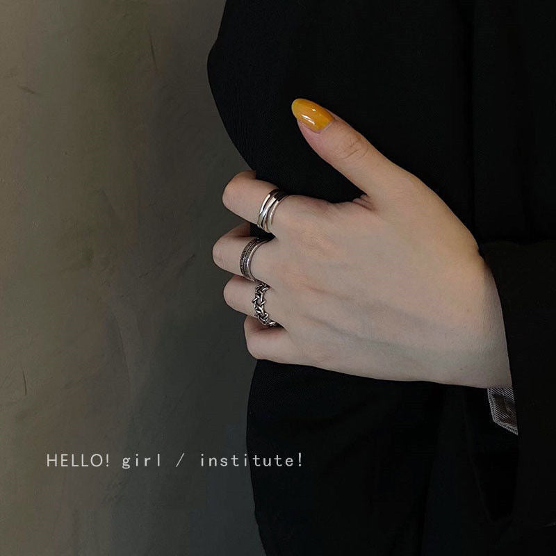 A woman's hand with yellow nail polish wearing three silver rings on different fingers against a black outfit and neutral wall