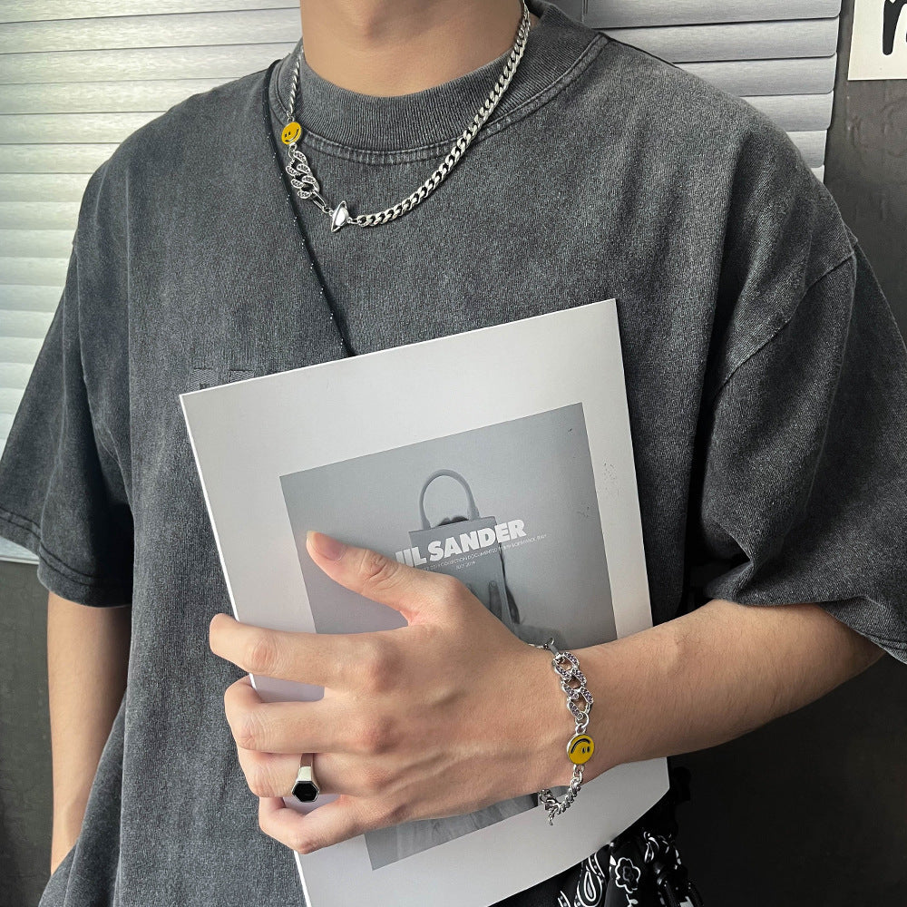 Titanium Steel Cuban chain necklace and matching bracelet with smiley face charm styled on model wearing gray t-shirt, holding Jil Sander book