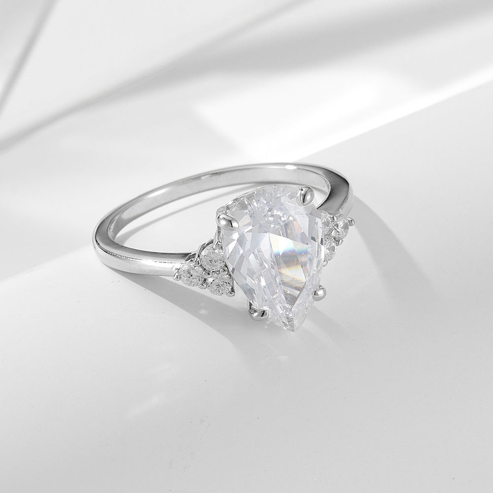 A silver engagement ring with pear-shaped diamond center and triangle accent stones, displayed on white fabric surface with soft shadows
