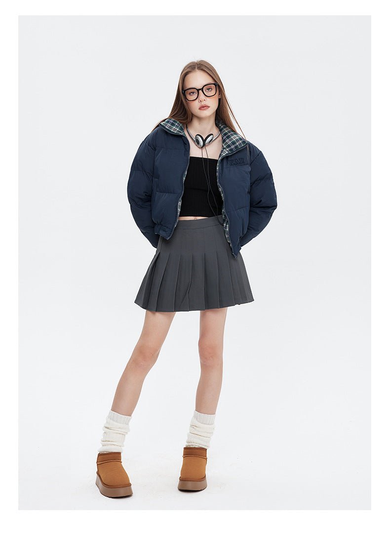 A woman wears a navy puffer jacket with a plaid collar, a black crop top, a gray pleated skirt, and brown platform boots