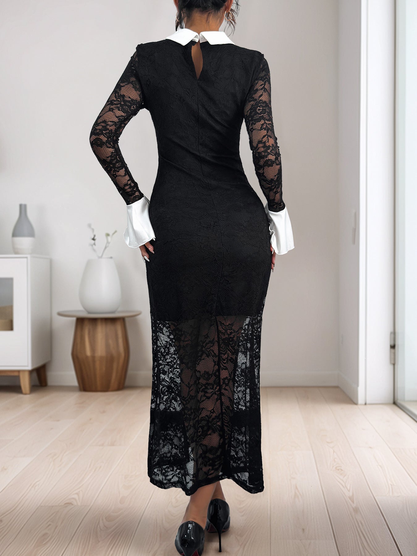 A woman showcasing the back of a black lace dress with a keyhole closure, white collar, and flared cuffs, styled with black heels.