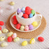 A white teacup filled with red, yellow, purple, and speckled mini eggs, placed on a saucer with scattered eggs around.