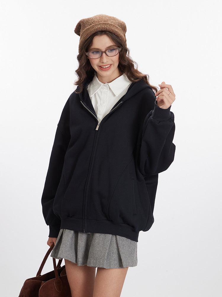 A model wears a black oversized hoodie, white shirt, and gray pleated skirt, holding a brown suede handbag while smiling
