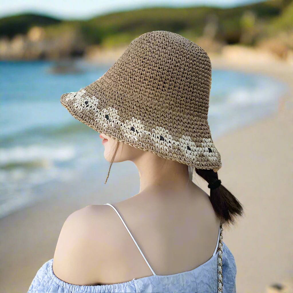 A young woman seen from the back, wearing a brown woven bucket hat with a floral pattern, styled with a blue off-shoulder dress and a small chain bag.