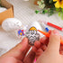 A person coloring a white Easter egg with a chick design using an orange marker, with other decorated eggs and markers in the background.