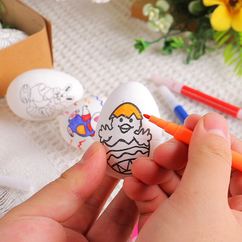 A person coloring a white Easter egg with a chick design using an orange marker, with other decorated eggs and markers in the background.