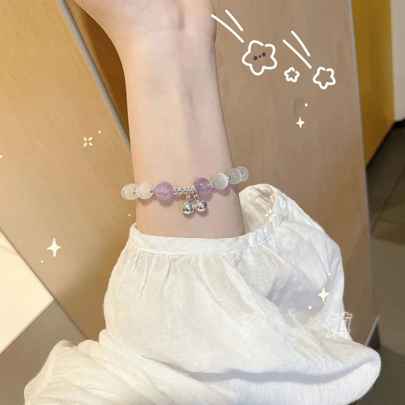 A woman wearing a white linen blouse showcasing a crystal bracelet with white and purple gemstones on her wrist. Small silver bell charms hang from bracelet