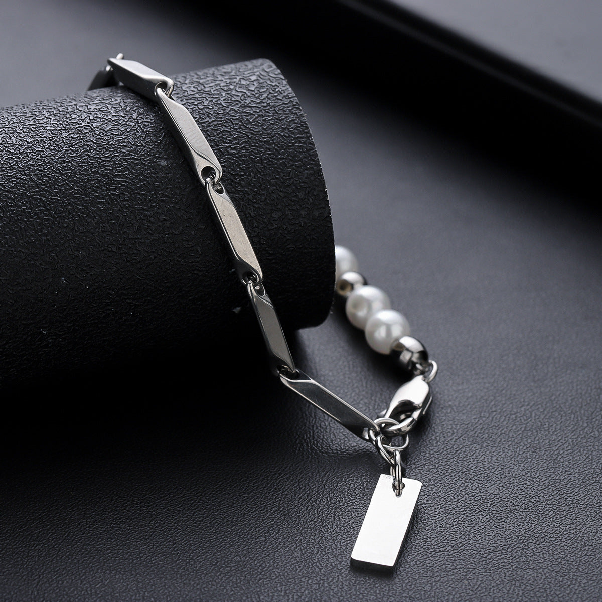 Silver chain bracelet with pearl accents and a rectangular tag charm, displayed on black textured surface. Sleek and stylish accessory.