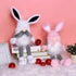 White and pink plush Easter gnome figurines with dangling legs, fluffy fur, and cute bunny ears, set against a pastel pink background.
