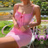 A woman wearing pink ribbed mini dress with tie detail sits in white garden chair surrounded by colorful flowers