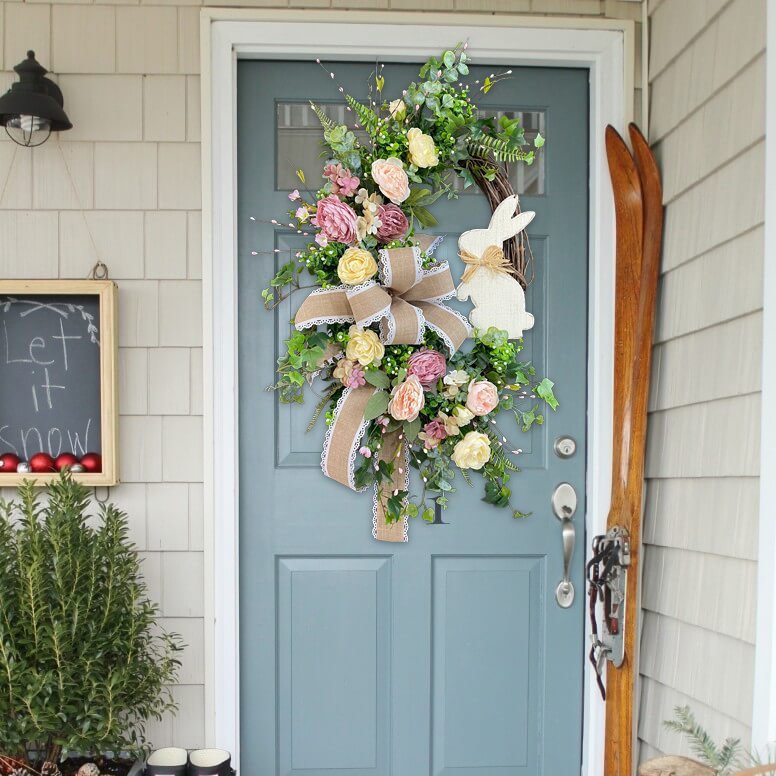 Easter wreath with pink, yellow, and cream flowers, burlap bow, and a wooden bunny, displayed on a light blue front door.