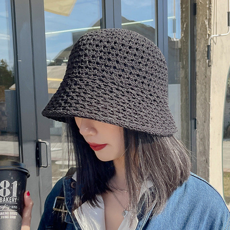 A woman in a black open-weave straw bucket hat, holding a coffee cup while wearing a denim jacket, standing outside a modern cafe.