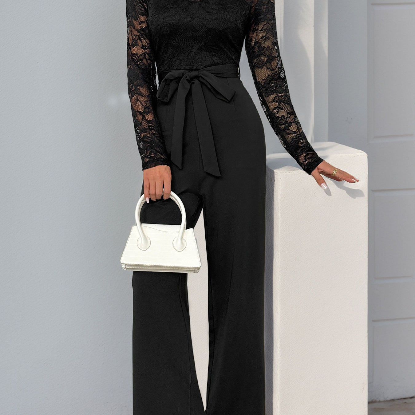 A woman wearing a black lace long-sleeve jumpsuit with a belted waist, paired with white heels and a white handbag, posing against a white wall.