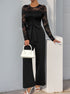A woman wearing a black lace long-sleeve jumpsuit with a belted waist, paired with white heels and a white handbag, posing against a white wall.