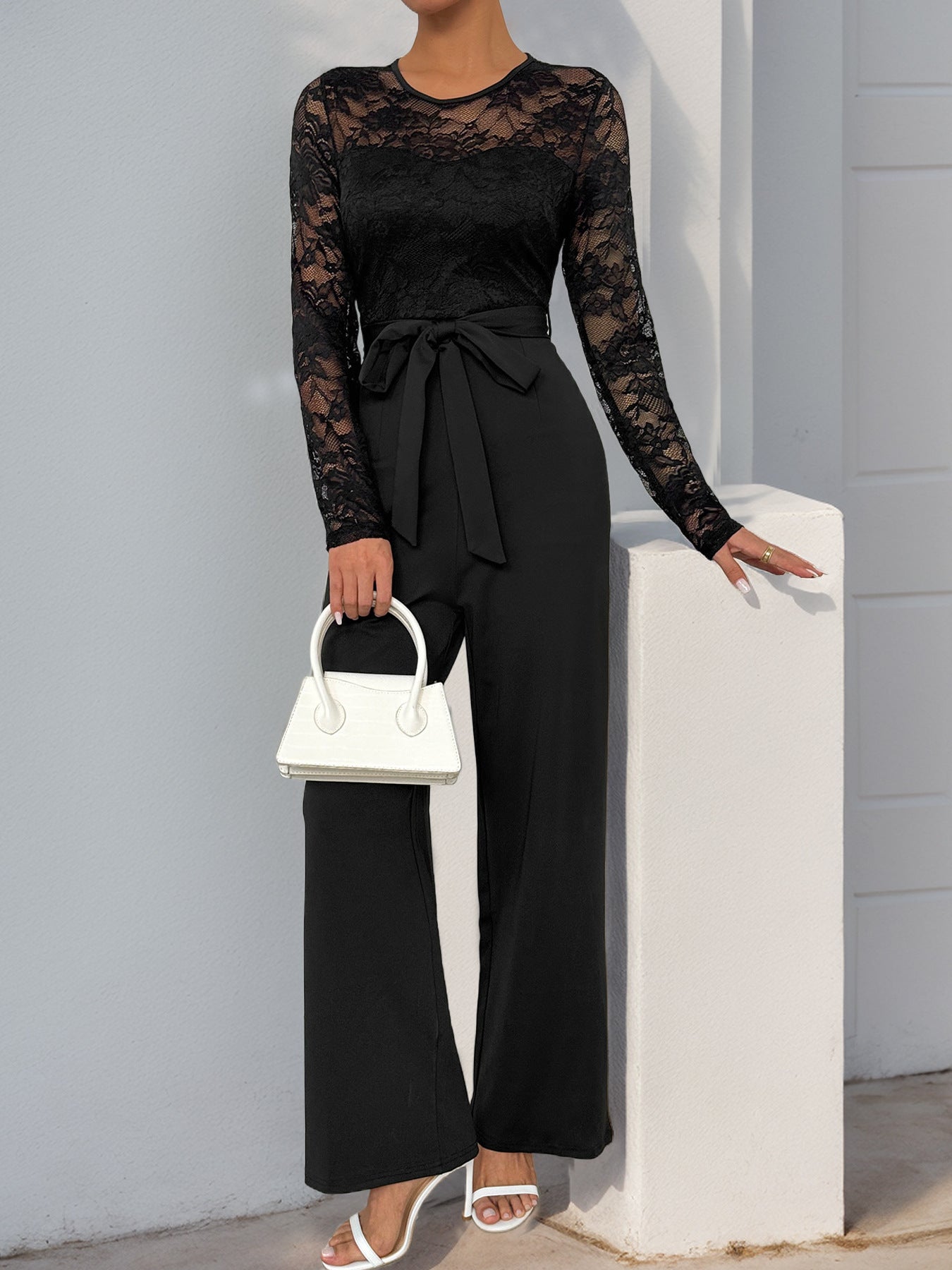 A woman wearing a black lace long-sleeve jumpsuit with a belted waist, paired with white heels and a white handbag, posing against a white wall.
