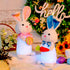  Side view of two glowing Easter bunny plush toys with soft fur, one in a pink scarf holding an egg and the other in a blue bowtie holding a carrot, surrounded by Easter decorations.
