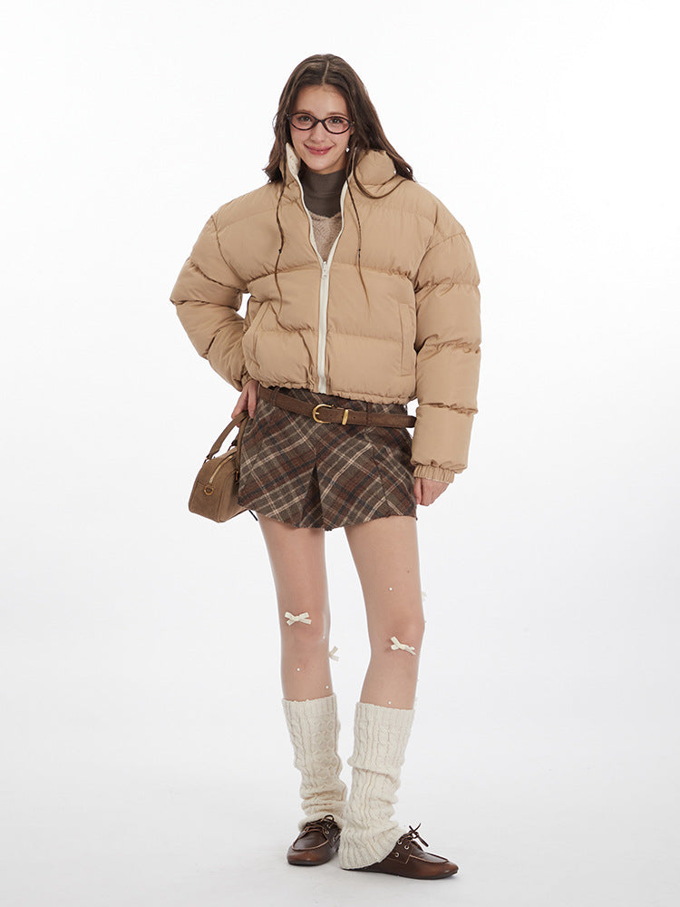 Full-body shot of a woman wearing a beige puffer jacket, plaid skirt, knee-high knit leg warmers, and brown loafers