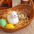 A white glittery chick figurine nestled in a wicker basket with yellow shredded paper, beside colorful Easter eggs and a nest with speckled eggs.