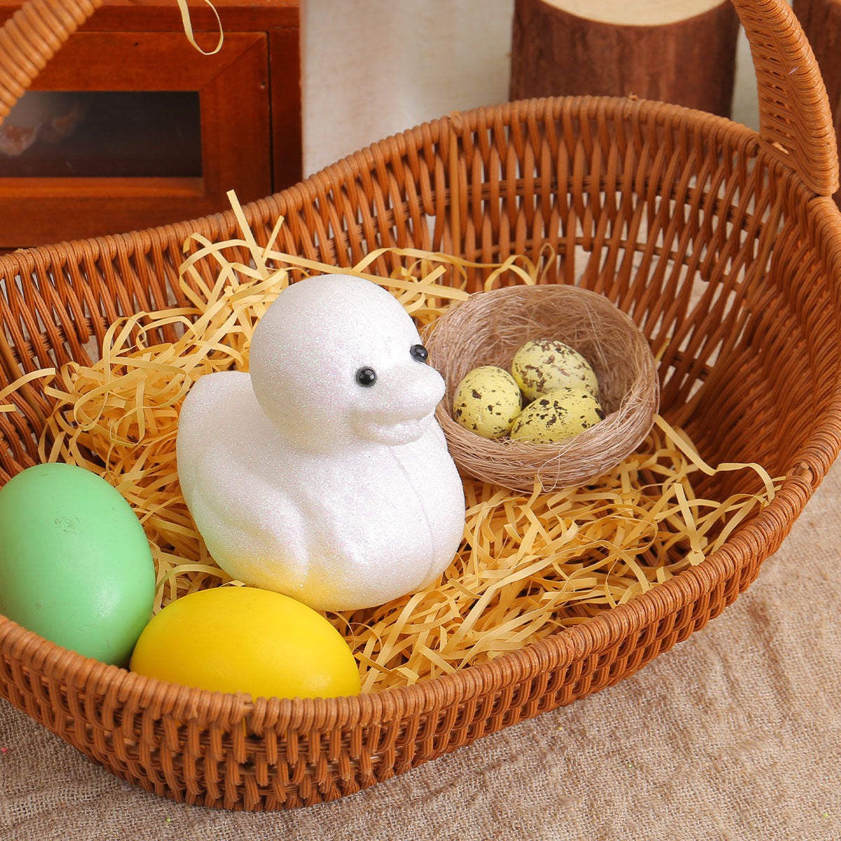 A white glittery chick figurine nestled in a wicker basket with yellow shredded paper, beside colorful Easter eggs and a nest with speckled eggs.