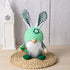 Green bunny gnome plush with a knit hat and carrot decoration, sitting on a woven mat with a neutral background.
