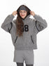 Close-up of grey textured hoodie highlighting '08' graphic appliqué, black whipstitch trim, and hood construction. Shown with grey cable knit shorts