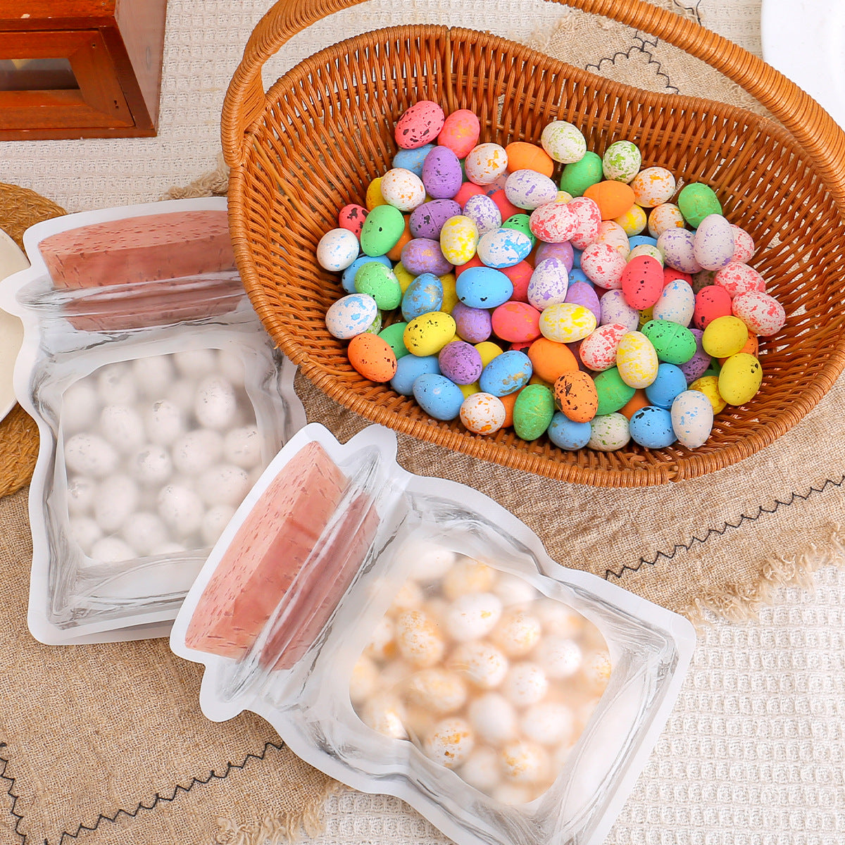 A woven basket filled with colorful Easter eggs, with two transparent jar-shaped bags containing white and golden-speckled eggs nearby.