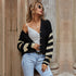 A woman leaning against stone wall wearing a black cardigan with cream stripes, white tank top, light denim jeans and round sunglasses with wavy blonde hair