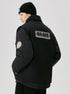Back view of black utility jacket showing BRAVE text patch and special number badge, styled with black beanie