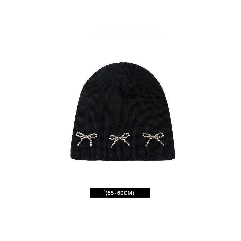 Black knit beanie with three pearl-studded bow decorations, with size measurement (55-60 cm) noted