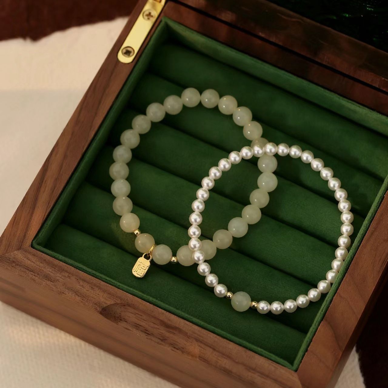 Two jade and pearl beaded bracelets displayed in a green-lined wooden jewelry box, featuring gold-toned details and small pendants