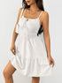 A woman is wearing a white cotton sundress with front tie detail and tiered skirt, carrying a small crossbody bag against a white backdrop