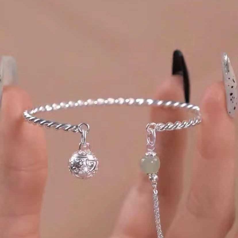 A silver twisted bracelet with decorative ball charm and jade bead pendant held between fingers