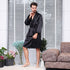 Man standing barefoot on white carpet wearing a black silk robe, hand thoughtfully touching his face against a backdrop of white blinds and decorative plants