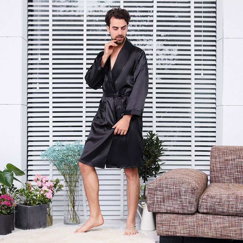 Man standing barefoot on white carpet wearing a black silk robe, hand thoughtfully touching his face against a backdrop of white blinds and decorative plants