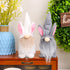 White and gray gnome plush toys with long beards, positioned on a wooden cabinet with decorative plants and a blue vase.