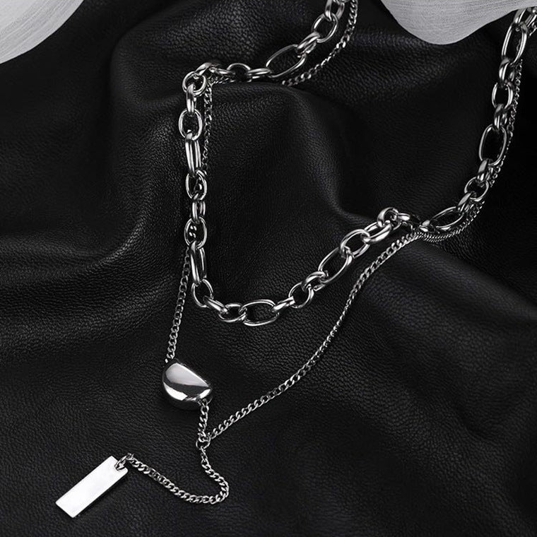 Multi-layer silver chain necklace with thick and thin chains, heart-shaped pendant, and rectangular tag on black leather background