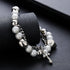 A silver bracelet with white beads, metallic accents, and a cross pendant wrapped around a black textured cylinder, highlighting its intricate details.
