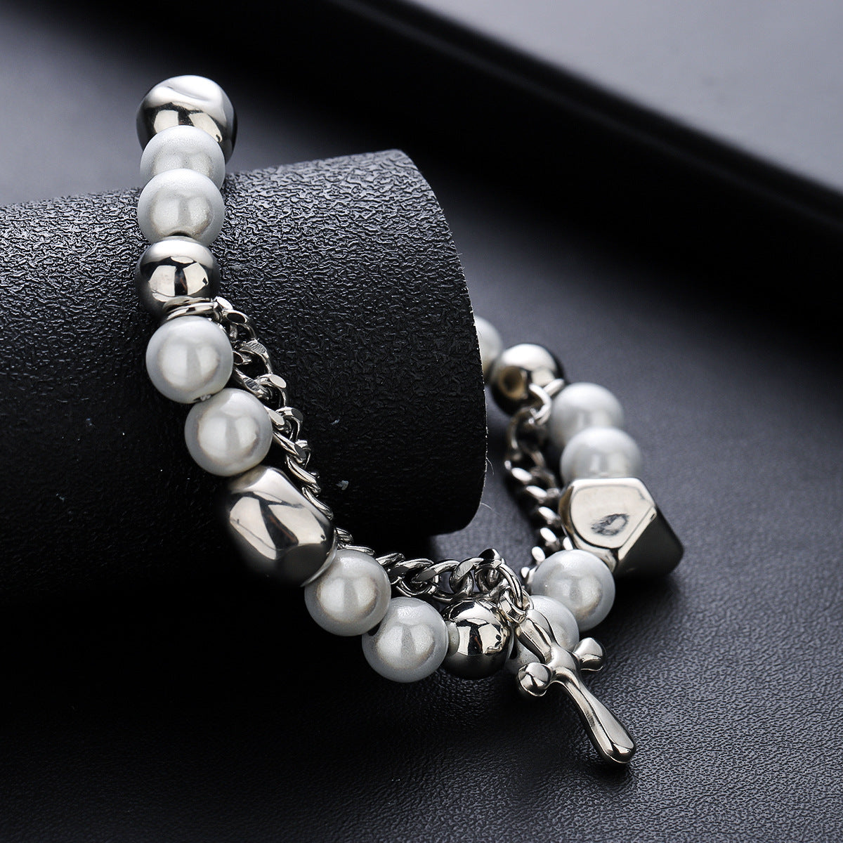 A silver bracelet with white beads, metallic accents, and a cross pendant wrapped around a black textured cylinder, highlighting its intricate details.